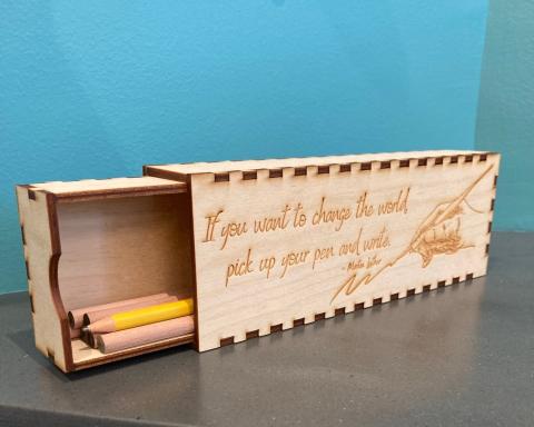 A photo of a sliding wooden box containing several wooden pencils. The box's edges are dark, and the joins between pieces are very visible. The box sits on a grey countertop with a blue wall behind it.